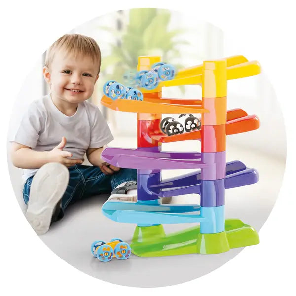 Smiling toddler with a colorful six-tier racing ramp and small cars flipping down vibrant zig-zag tracks.
