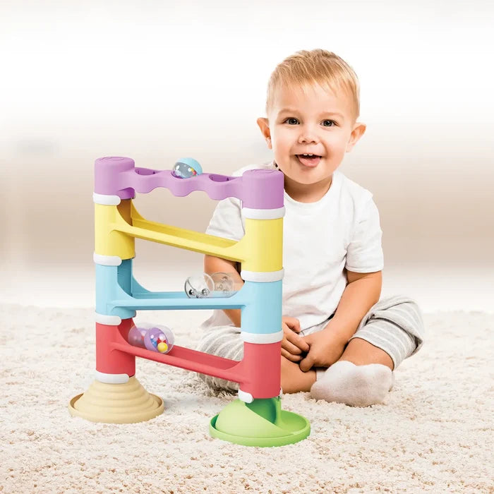 A toddler sits by a colorful four-tier pastel marble run tower with transparent rattle balls on a rug.