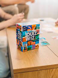 Colorful rectangular game box with mosaic illustrations and bold text on a wooden table with people playing.
