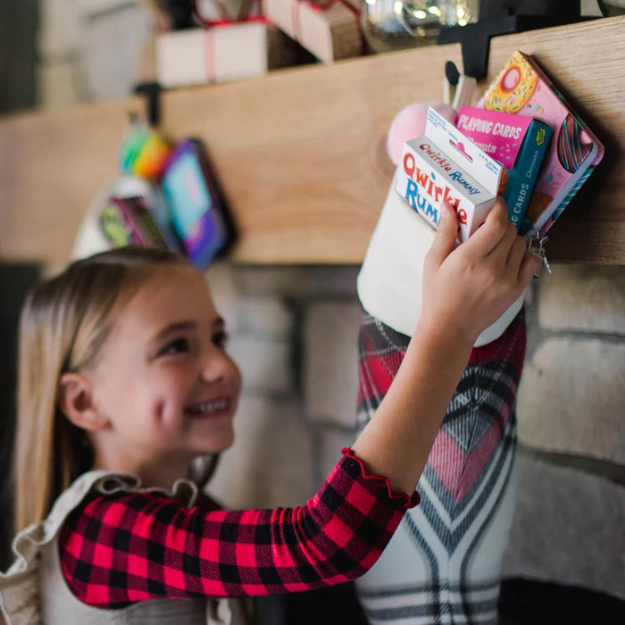 Girl pulls a small white Qwirkle Rummy box with red and blue lettering from a white stocking hung on a mantel.