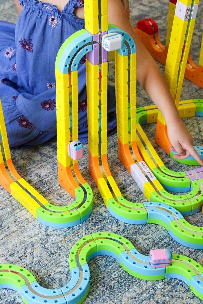 Vibrant yellow, green, and blue interlocking cube tracks forming towers and loops with small toy trains and a child.