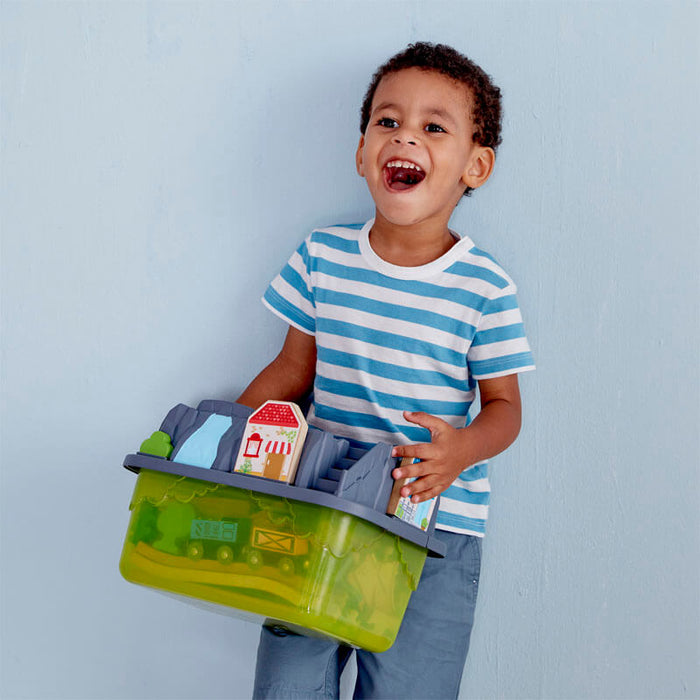 Smiling boy holding a green railway bucket with a grey mountain-top lid and small toy house.