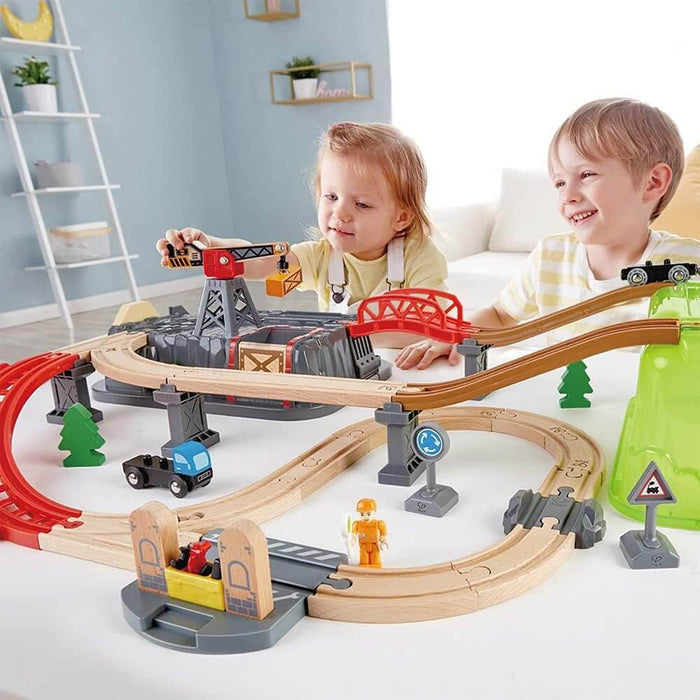 Two children play with a multi-level wooden train set featuring a red bridge, crane, and green mountain tunnel.