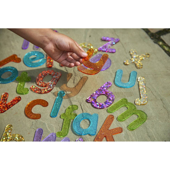 Hand picking up a translucent orange 'y' from colorful, glittery lowercase letters scattered on a stone surface.