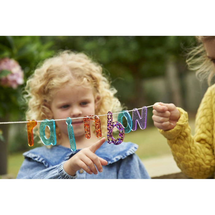 Child points to colorful translucent glittery letters spelling rainbow on a string in an outdoor garden.