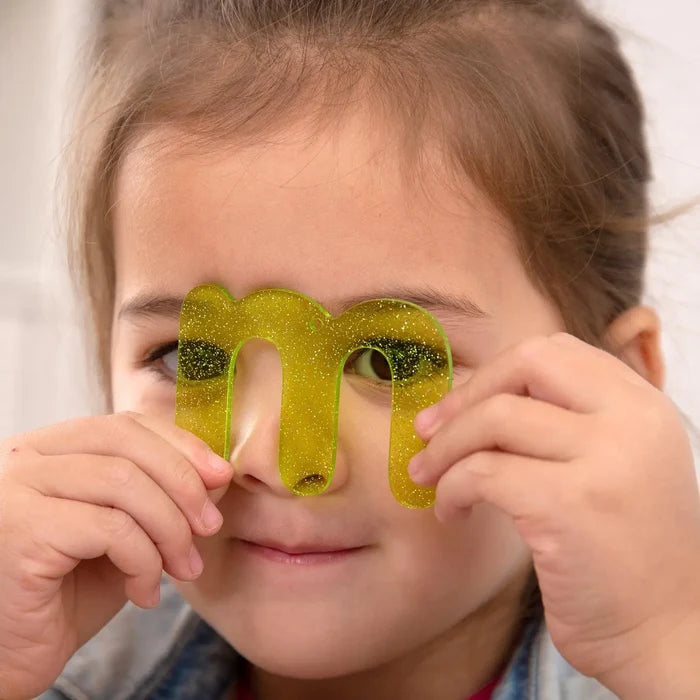 A child holding a translucent lime green glittery acrylic letter m in front of her eyes.
