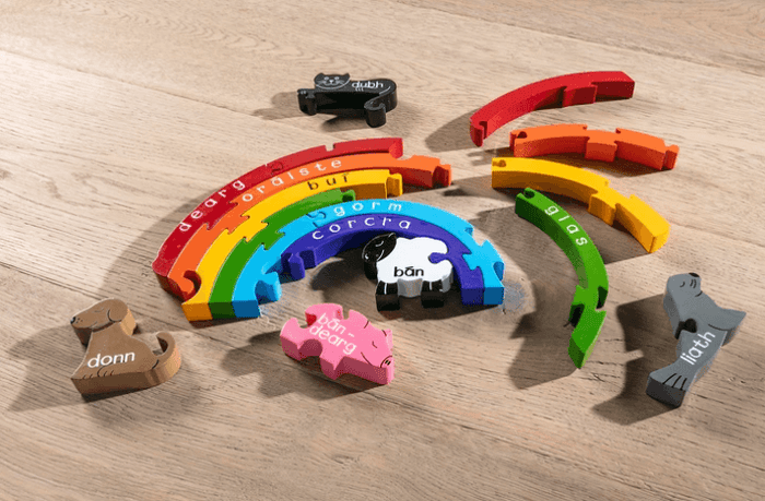 Colorful wooden rainbow arcs and animal shapes with Irish names like dearg and bán, on a light wood surface.