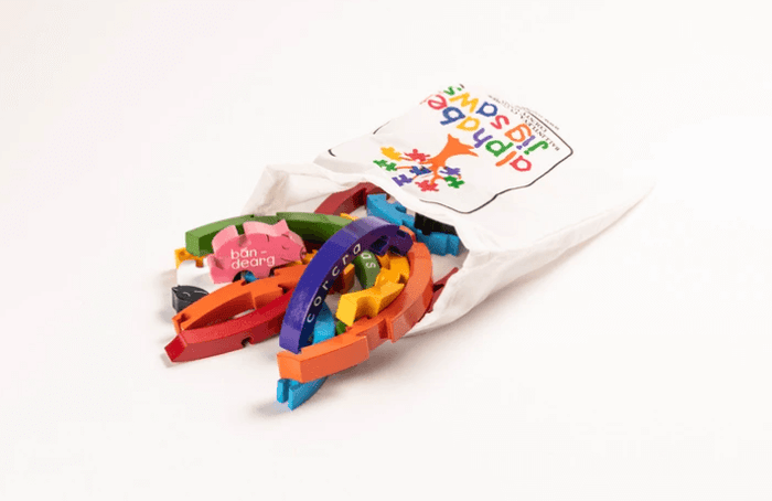 Vibrant arched wooden puzzle pieces with Irish words spill from a white drawstring bag with a colorful tree logo.