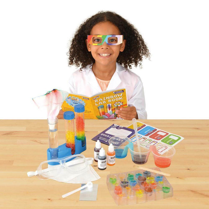 Child in lab coat and rainbow glasses with colorful test tubes, mixing tray, and science book on a wooden table.