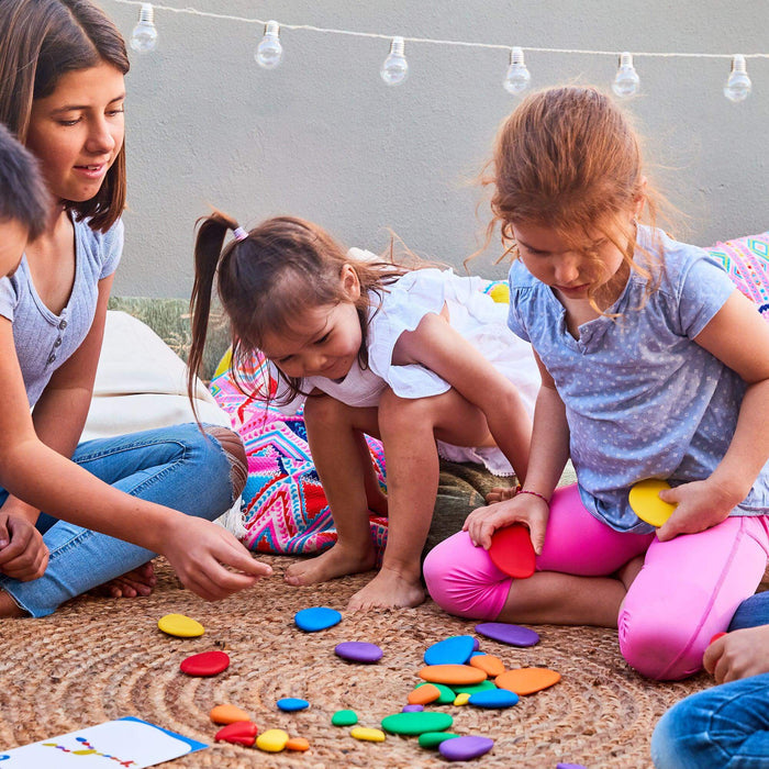 Three children play with smooth, colorful, oval-shaped stones in various sizes on a woven rug.