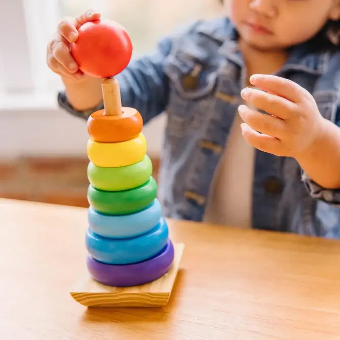 Child stacking a red wooden ball on top of a vertical tower of colorful rainbow rings on a wooden base.