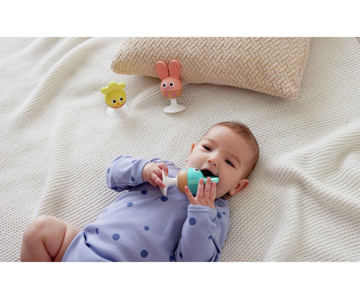 Baby lying on a white blanket holds a teal fish teether with a yellow chick and pink bunny teether standing nearby.