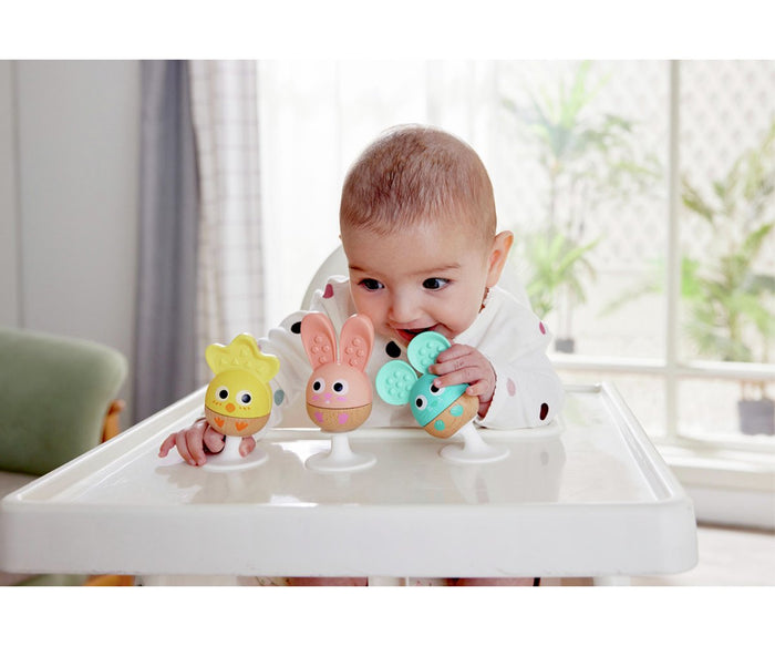 Baby playing with yellow chick, pink bunny, and teal mouse rattles with suction bases on a white high chair tray.