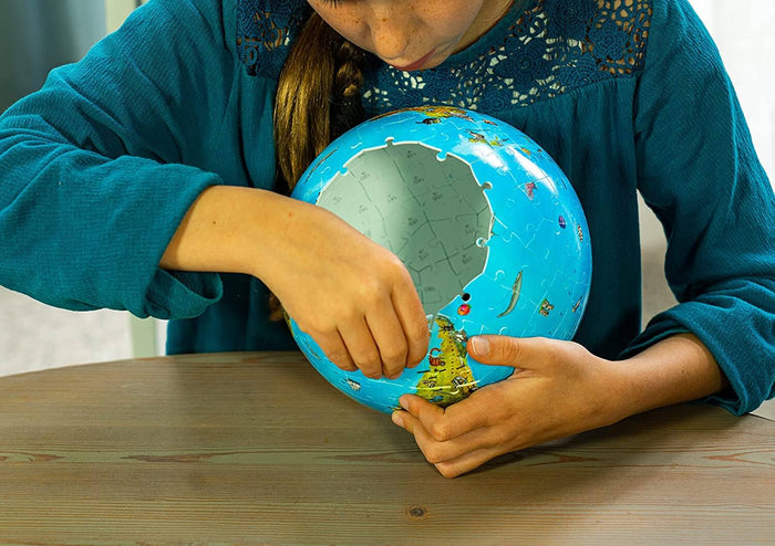 Child's hands assembling a light blue 3D globe jigsaw puzzle with animal illustrations on a wooden table.