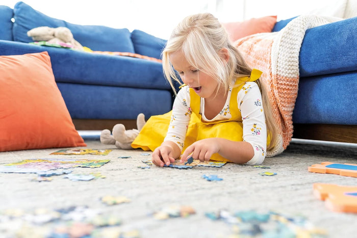 Young girl in a yellow dress assembles colorful jigsaw puzzles on a carpeted floor next to a blue sofa.