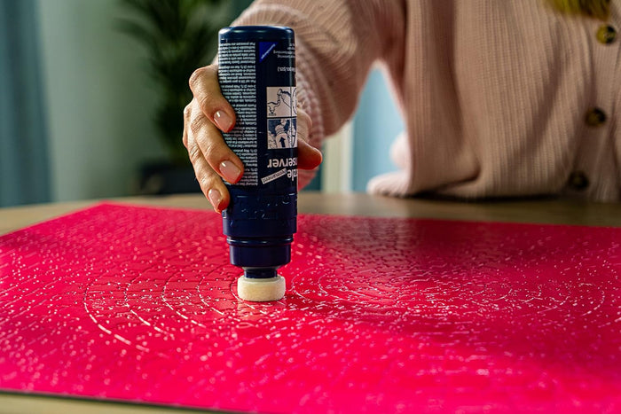 Hand applying clear glue from a blue sponge-tip bottle onto a bright pink jigsaw puzzle.