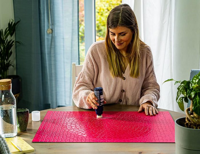 Woman applying puzzle glue with a blue sponge-tip bottle to a completed pink puzzle on a wooden table.