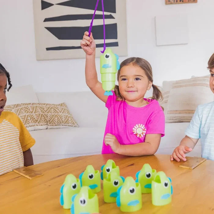 A girl catches a green plastic fish with a purple rod. Several more green fish sit on a table as children play.