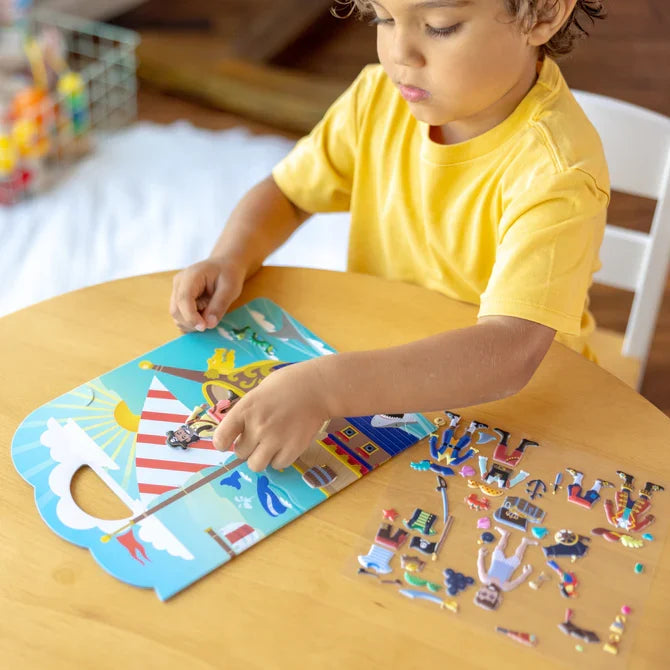 Child in a yellow shirt places pirate puffy stickers on a bright blue sea scene board with a pirate ship.