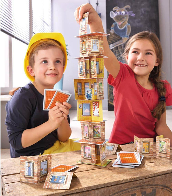 Two children building a tall tower using illustrated brick-patterned folding cards and flat cards on a wooden table.