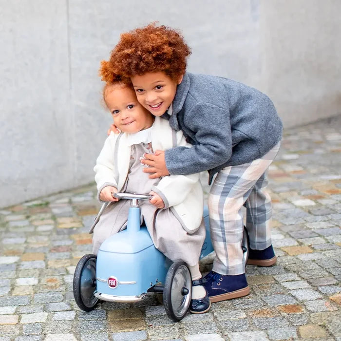 Light blue Fiat 500 ride-on toy car with chrome accents, shown with two children on a cobblestone street.