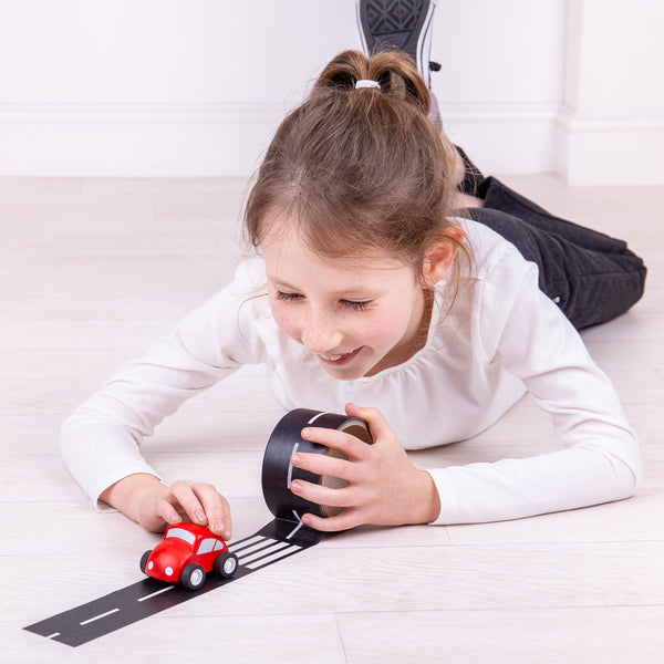 Girl unrolling black tape with white road markings on a light floor with a small red toy car.