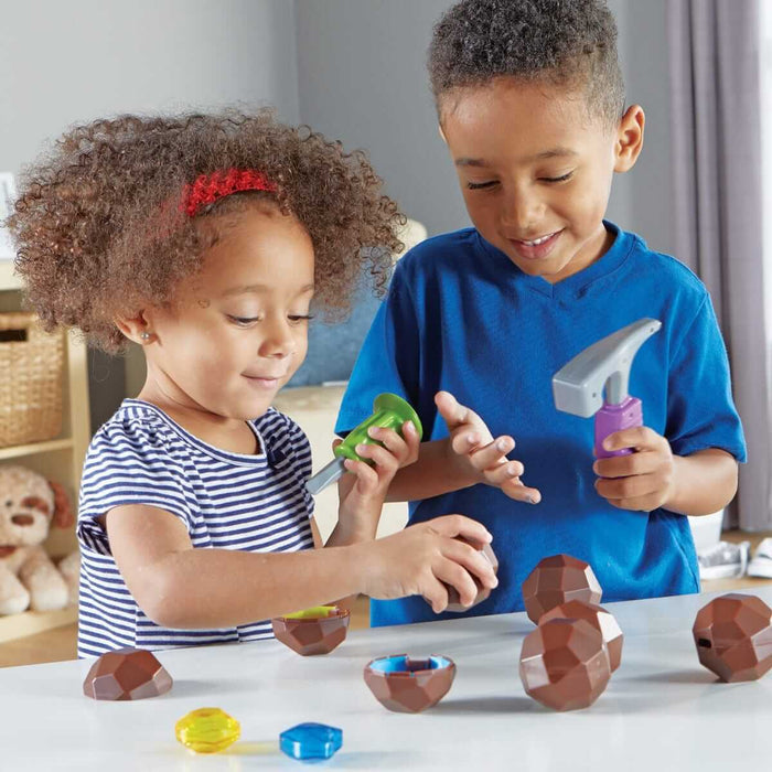 Two children using toy tools to crack open faceted brown rocks and discover colorful hidden gems.