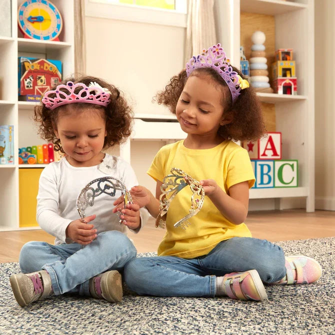 Two girls wearing pink and purple tiaras and holding silver and gold ones while sitting on a rug in a playroom.
