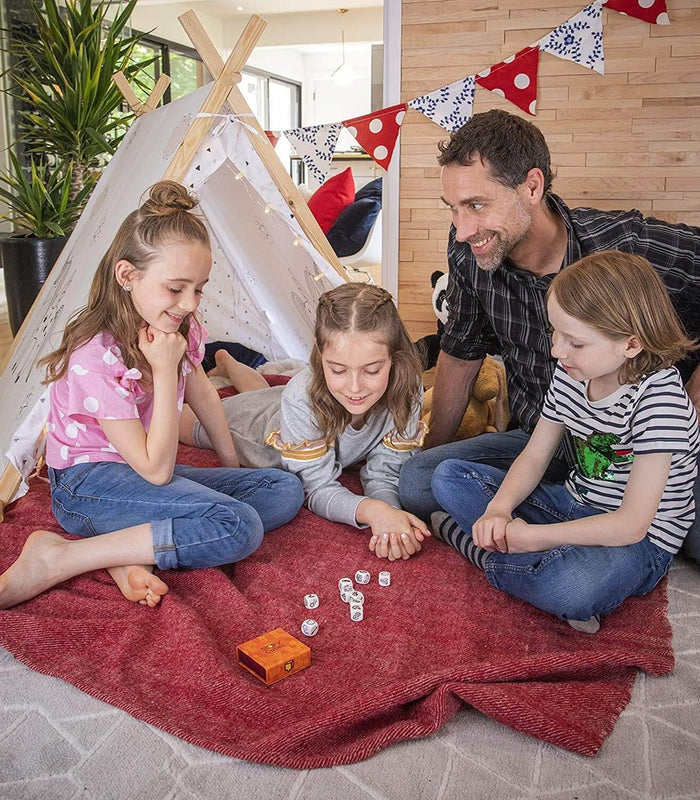 Family playing with white symbol dice on a red blanket near an orange box and a white indoor teepee.