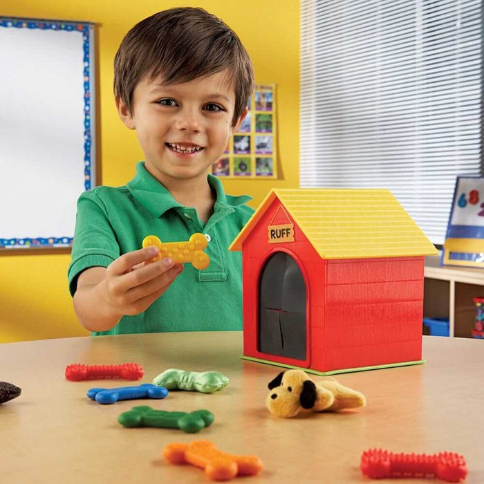 A boy holds a yellow textured bone by a red toy doghouse with colorful bones and a plush dog on a table.