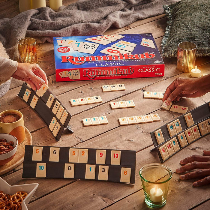 Hands playing Rummikub with numbered tiles in black racks on a wooden table with the blue game box and cozy snacks.