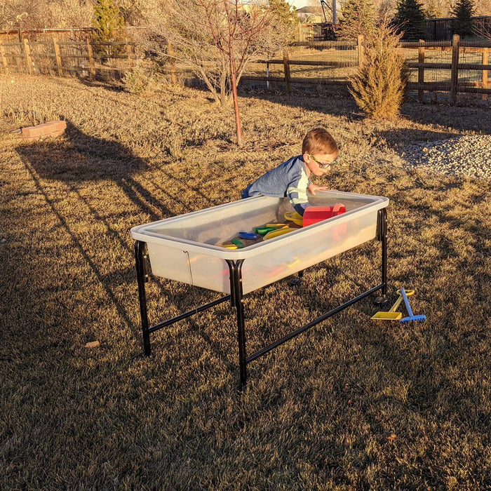 Child playing at a clear rectangular tray on a black metal stand, filled with sand and toys on a sunny lawn.
