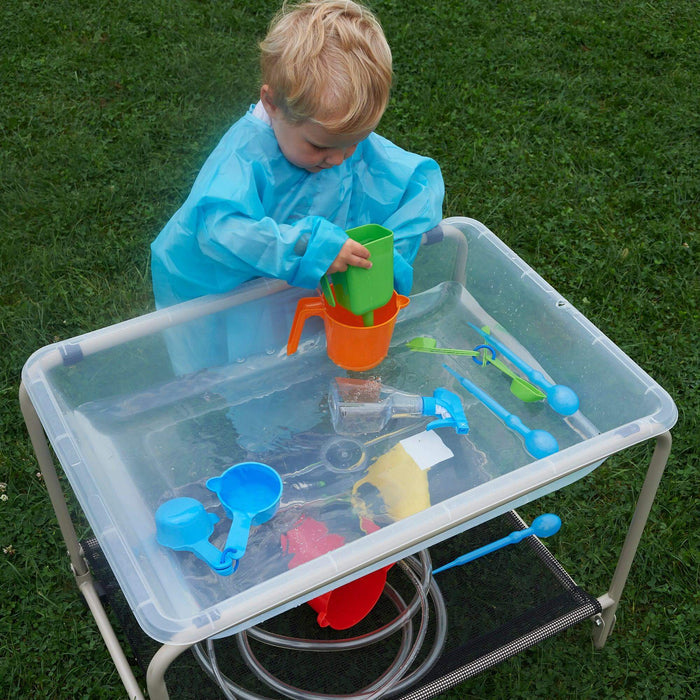 Young child playing with colorful scoops and pitchers in a clear water table on a metal stand, outdoors on grass.