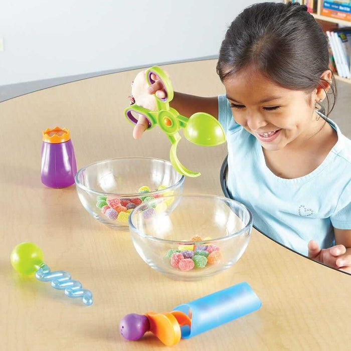 A child uses green scoop tweezers and colorful plastic droppers to move treats between two clear bowls.