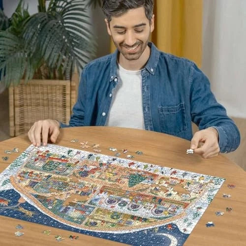 Smiling man in a blue shirt assembles a large, colorful festive jigsaw puzzle on a round wooden table at home.
