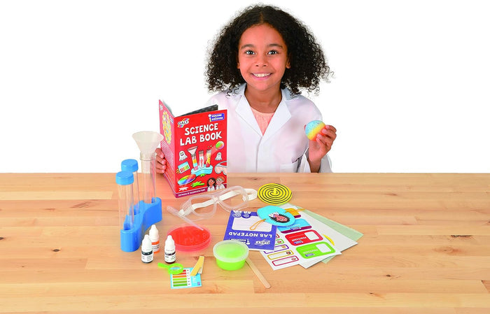 Girl in a white lab coat with Galt Science Lab kit: test tubes, goggles, red book, and slime on a wooden table.