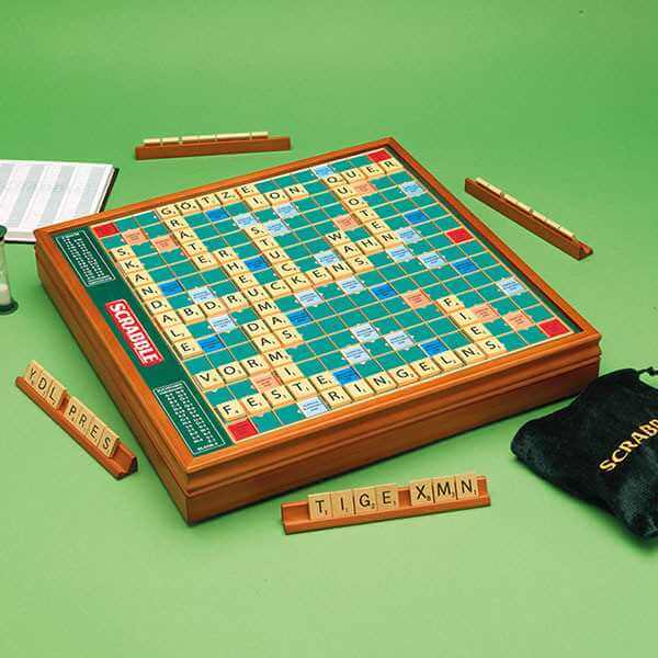 Elevated wooden Scrabble board with matching tile racks, wooden tiles, and a black velvet bag on a green background.