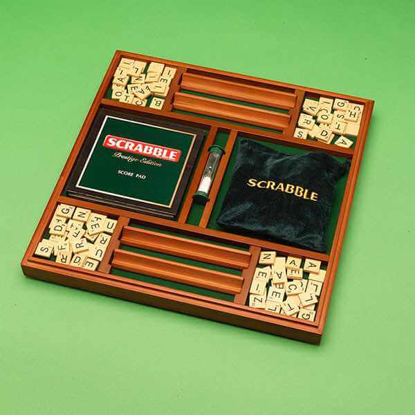 Wooden tray containing Scrabble letter tiles, four racks, a green score pad, sand timer, and a black velvet bag.