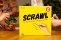 Yellow square box with black SCRAWL text and a hand drawing a suggestive line, shown with people playing at a table.