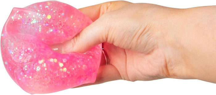 A hand squeezes a sparkly, translucent pink squishy cube toy filled with multi-colored iridescent glitter.