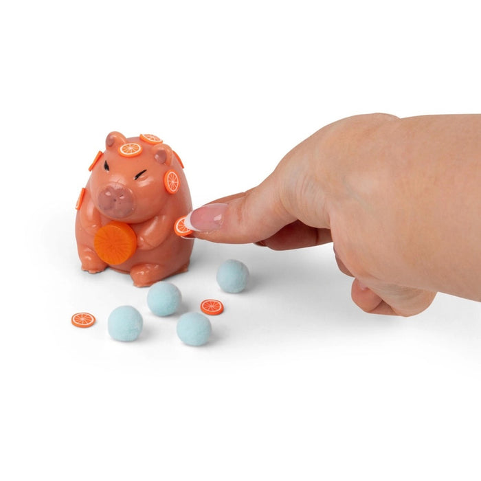 Hand decorating a peach squishy capybara toy with tiny orange slices, next to four blue fuzzy pom-poms.