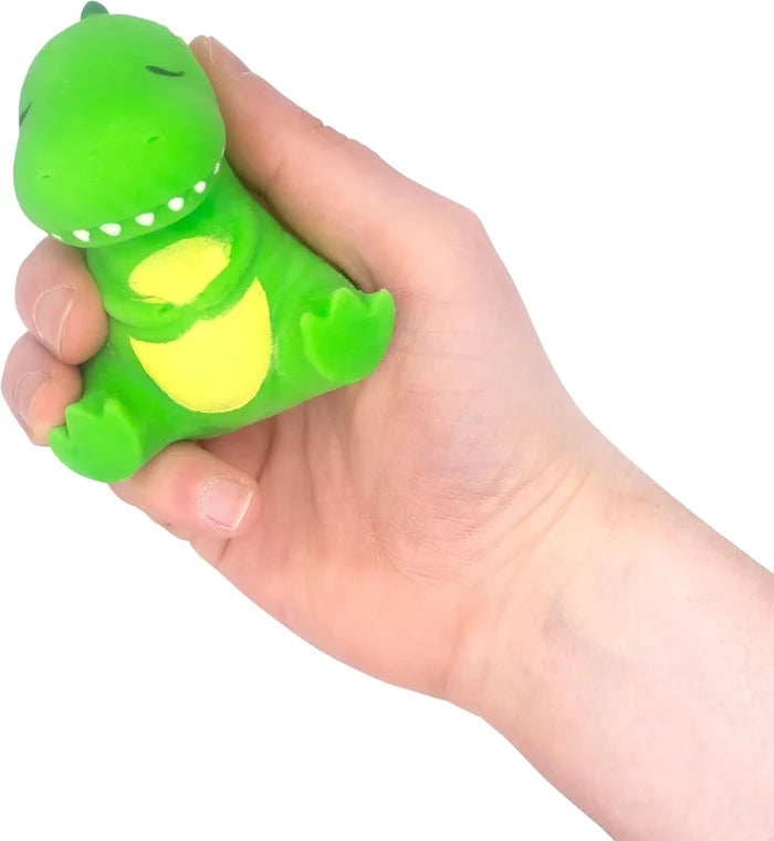 A hand squeezes a small green squishy dinosaur toy with sleepy closed eyes, white teeth, and a yellow belly.