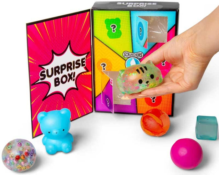 Hand pulling a cat squishy from a colorful surprise box with a blue bear and sensory toys in the foreground.