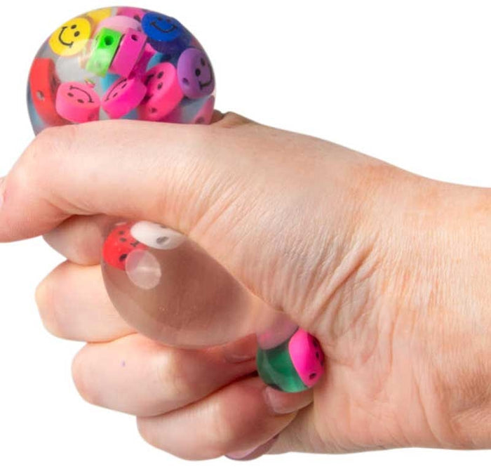 A hand squeezing a clear squishy ball filled with vibrant, multi-colored smiley face beads.