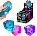 Hand squeezing a blue squishy cube beside pink and purple textured cubes and a Scrunchems retail display box.