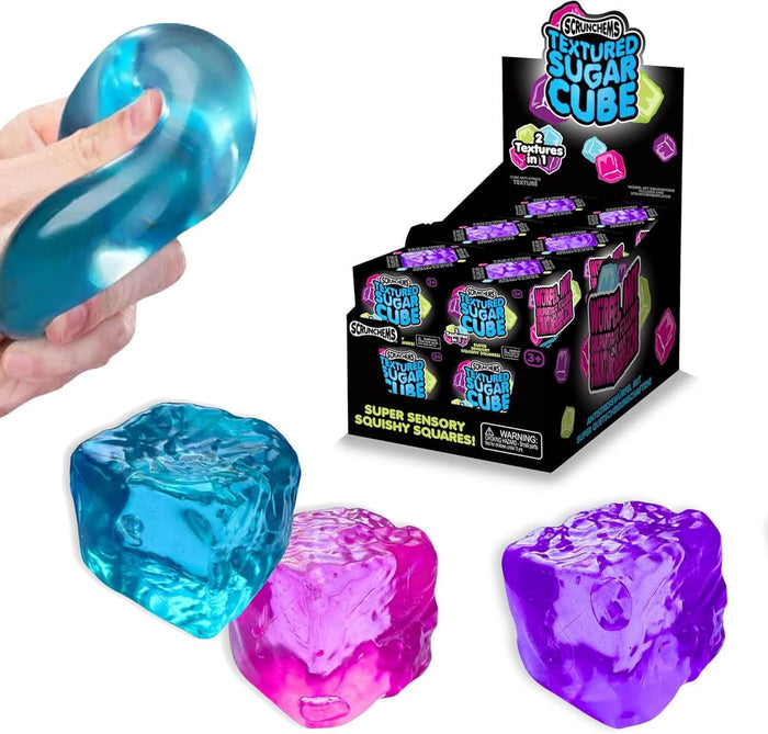 Hand squeezing a blue squishy cube beside pink and purple textured cubes and a Scrunchems retail display box.