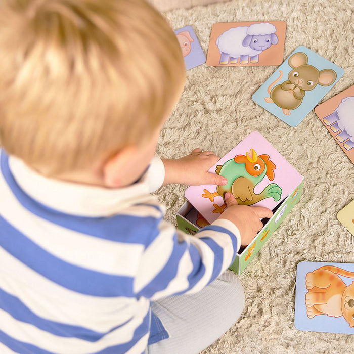 Toddler in a striped shirt puts a rooster card into a green box. Large animal cards lie on a shaggy rug.