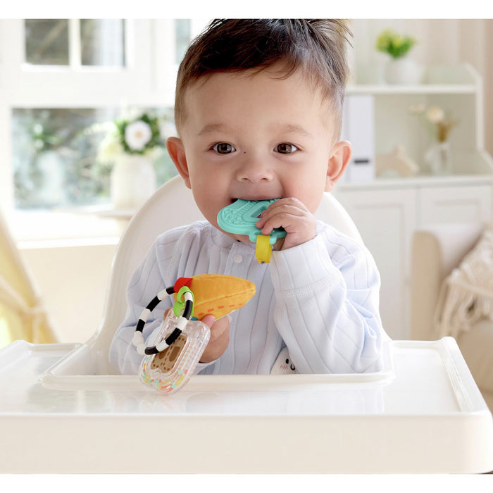 Baby in a high chair chewing on a teal textured toy and holding a plush orange slice and ring rattle.