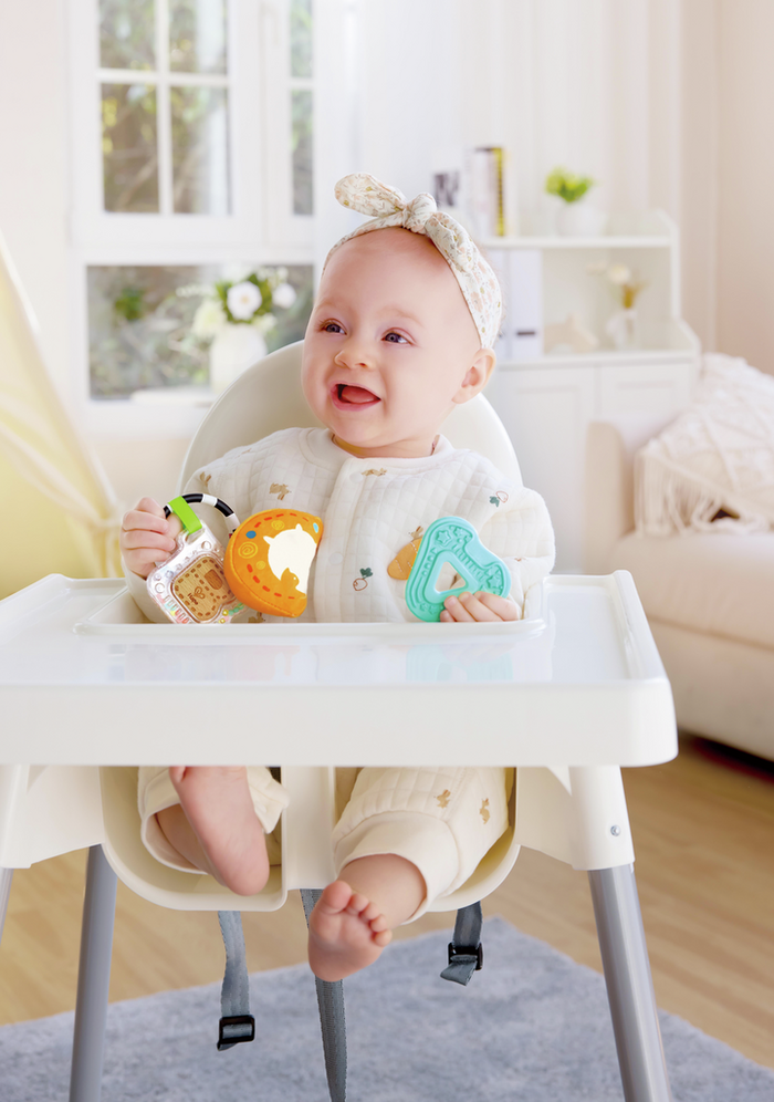 Baby in a high chair holding a teal letter A teether, orange mirror toy, and a clear rattle with colorful beads.