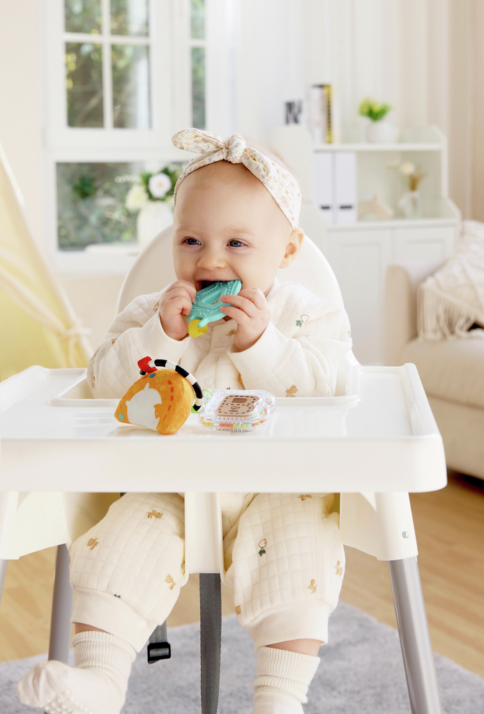 Baby in a high chair holding a teal teether, with an orange fabric toy and clear ABC rattle on the tray.